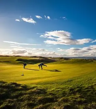 Golfers reading the green on a dramatic links course overlooking the sea near St Andrews, Scotland, under a vivid blue sky