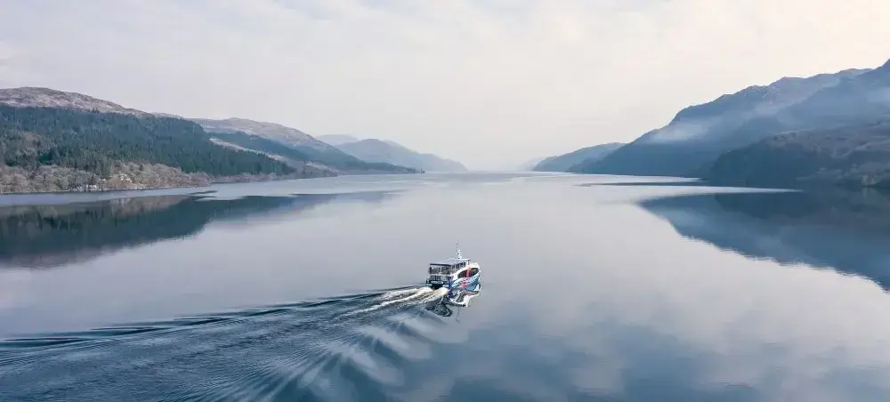 Aerial view of a private charter boat cruising across a glassy Scottish Highland loch surrounded by mist-covered mountains