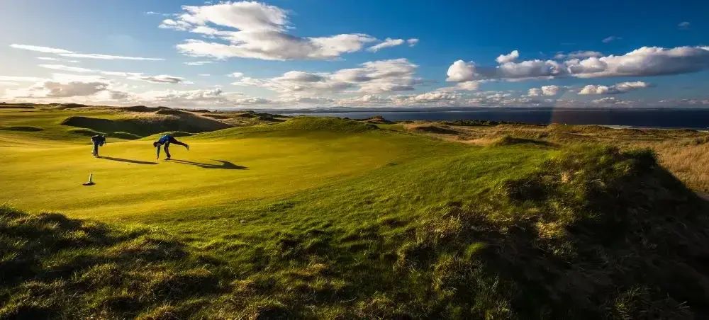 Golfers putting on a coastal links course in St Andrews, Scotland, with rolling dunes and ocean views under a dramatic sky