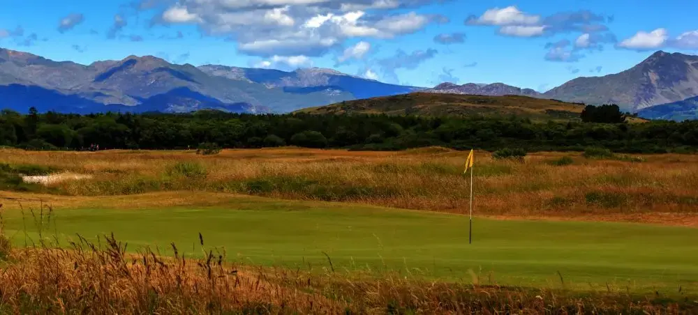 Links golf course with yellow flag amid wild rough grass, backed by the dramatic mountain peaks of Eryri (Snowdonia), Wales