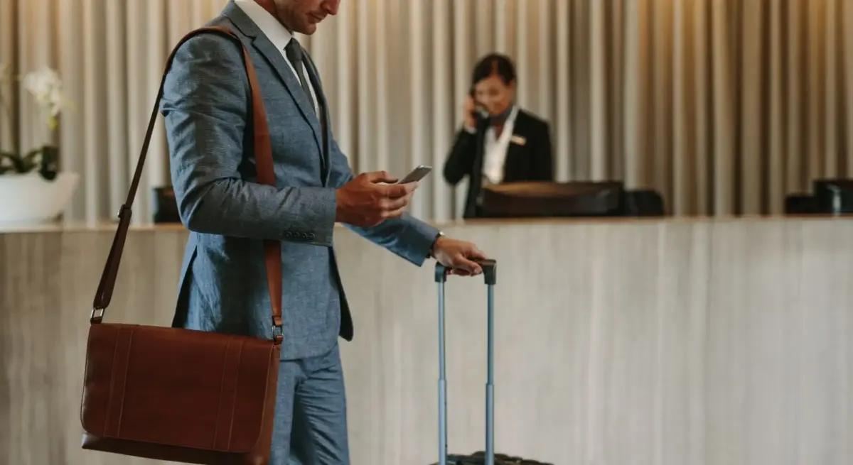 Business traveller in a tailored suit checking his phone at a luxury hotel reception, luggage in hand, booking a private jet charter for short-haul travel