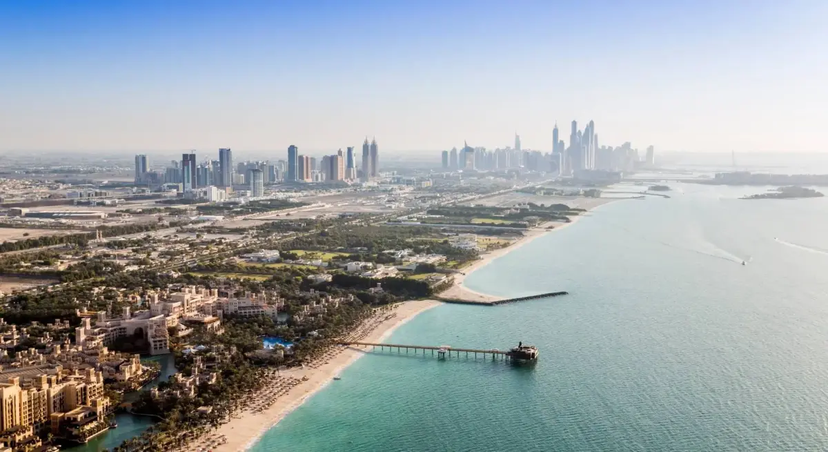 Aerial view of Dubai's luxury beachfront resorts and marina pier stretching into turquoise Arabian Gulf waters with the city skyline beyond