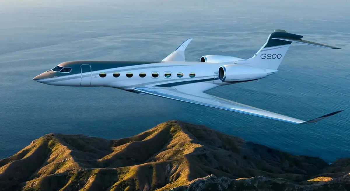 Gulfstream G800 ultra-long-range private jet cruising at altitude over coastal mountains, showcasing its sleek white exterior and signature oval windows