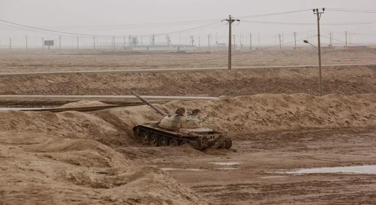Abandoned military tank partially buried in desert sand near a Middle Eastern roadway, illustrating ground-level conflict risks in the region