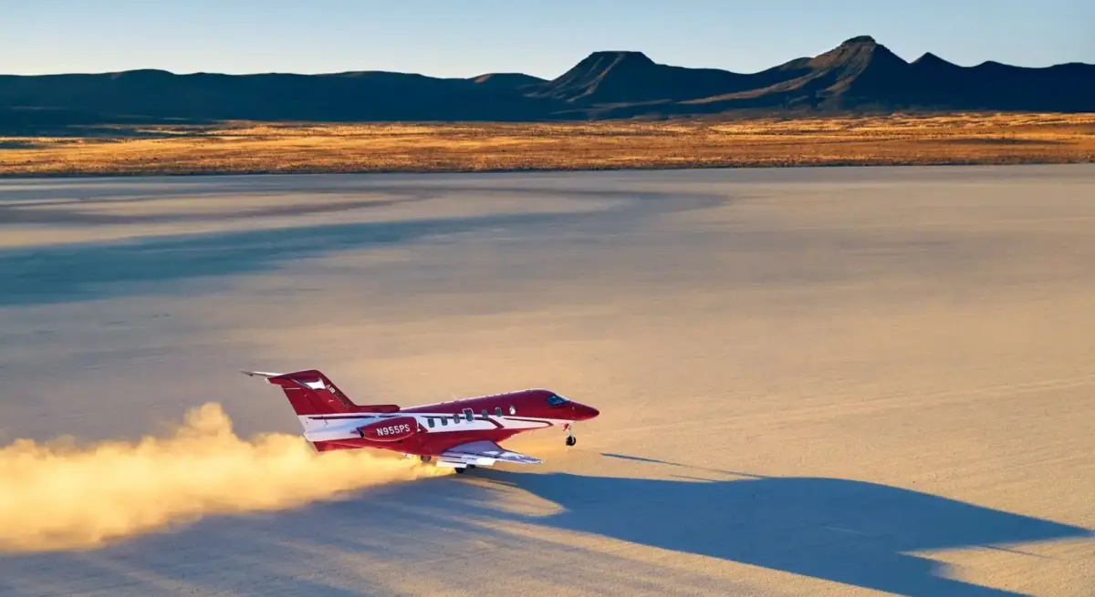 Red Pilatus PC-24 private jet taking off from a desert, trailing dust against a dramatic mountain backdrop at golden hour
