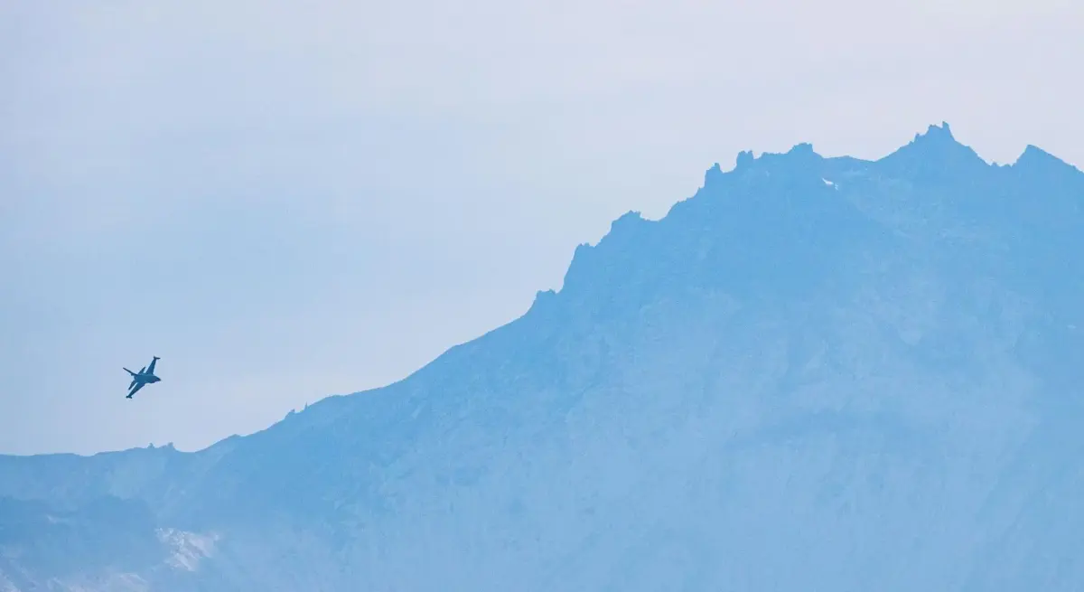 Small jet aircraft flying past a dramatic mountain ridge under a hazy blue sky, evoking unrestricted private aviation freedom